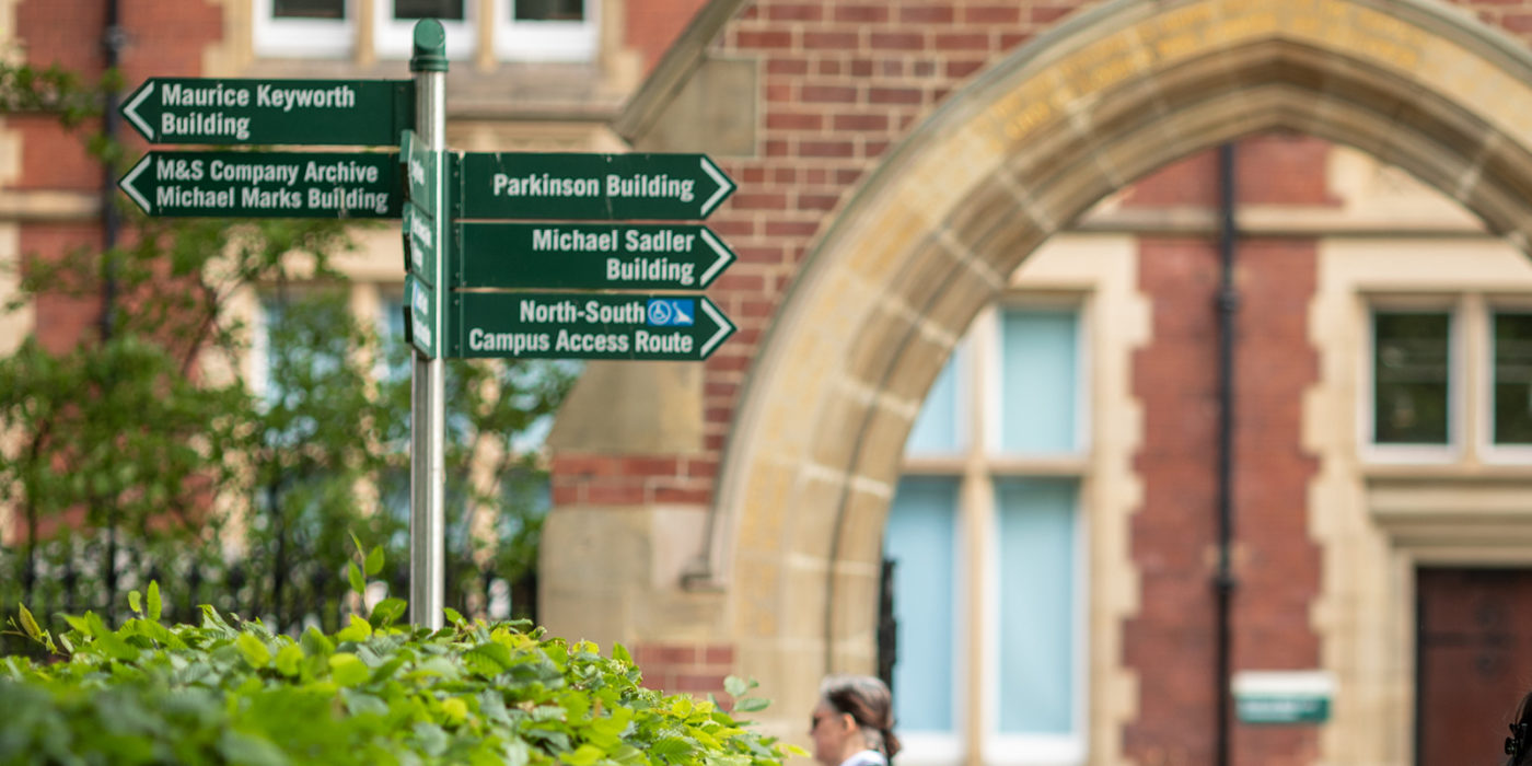 A signpost showing signs pointing to different University buildings in front of an old red brick building on campus.